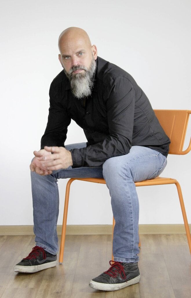 Moderner Mann sitzt auf orangefarbenem Stuhl vor weißem Hintergrund, casual Kleidung und nachdenkliche Haltung.